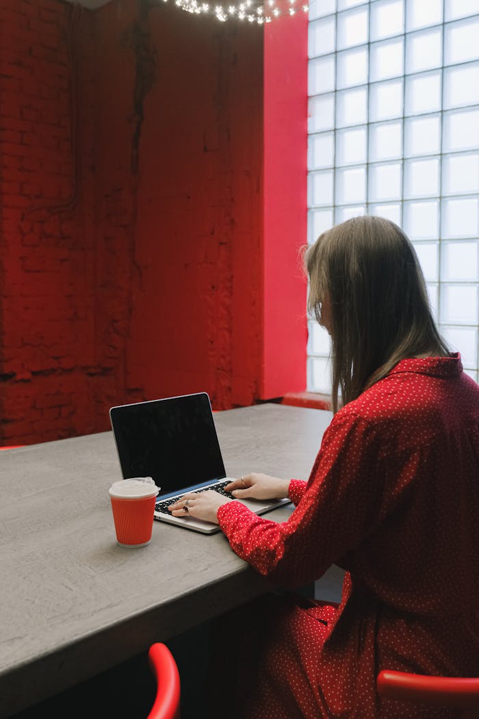 Woman in a red dress working on a laptop at a desk with a coffee cup indoors.